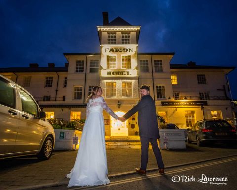 A bride and groom hold hands in front of a lit historical building at dusk.